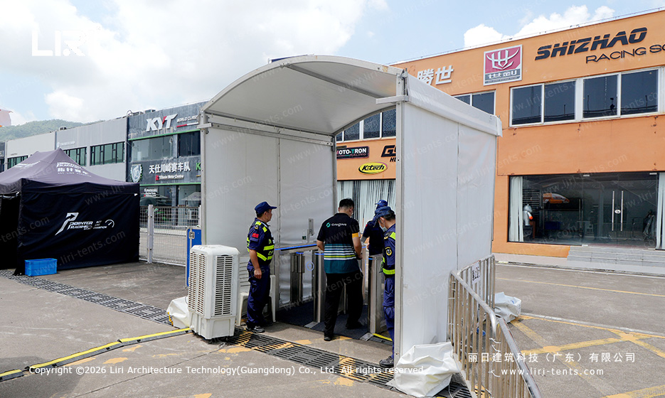 Curve roof A-frame security check entry tent at a race track pit lane with staff scanning credentials between two team pit buildings