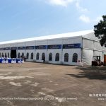 Wide exterior view of a long clear span A-frame event tent used as a spectator lounge at a race track, with arched glass windows and sponsor banners along the side