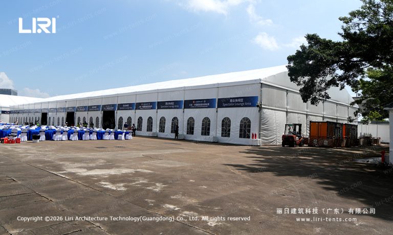 Wide exterior view of a long clear span A-frame event tent used as a spectator lounge at a race track, with arched glass windows and sponsor banners along the side