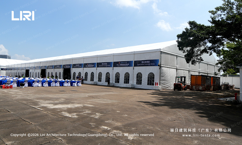 Wide exterior view of a long clear span A-frame event tent used as a spectator lounge at a race track, with arched glass windows and sponsor banners along the side