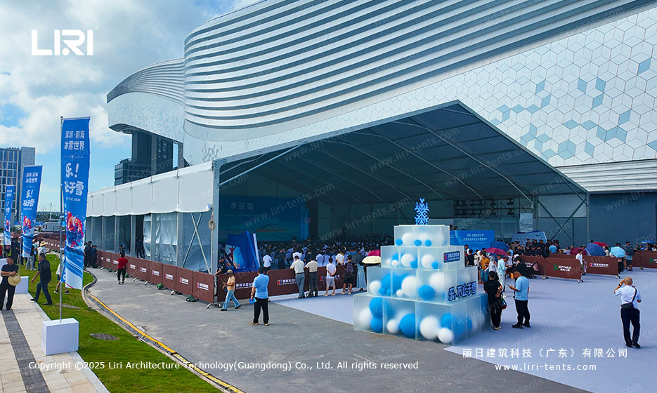 Side exterior of a clear span event tent with blue themed decorative cubes and crowds queuing at the entrance